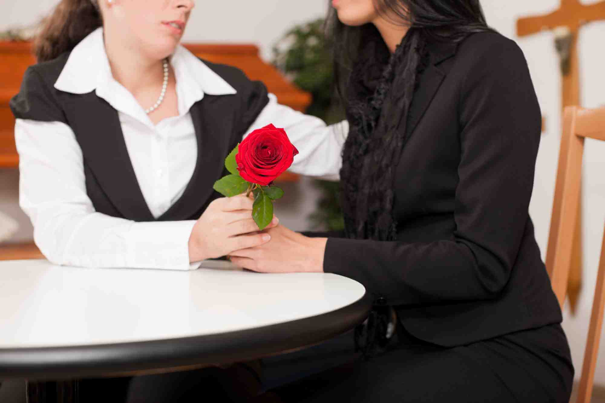 Female funeral director in formal clothes comforts mourning woman in black holding a red rose