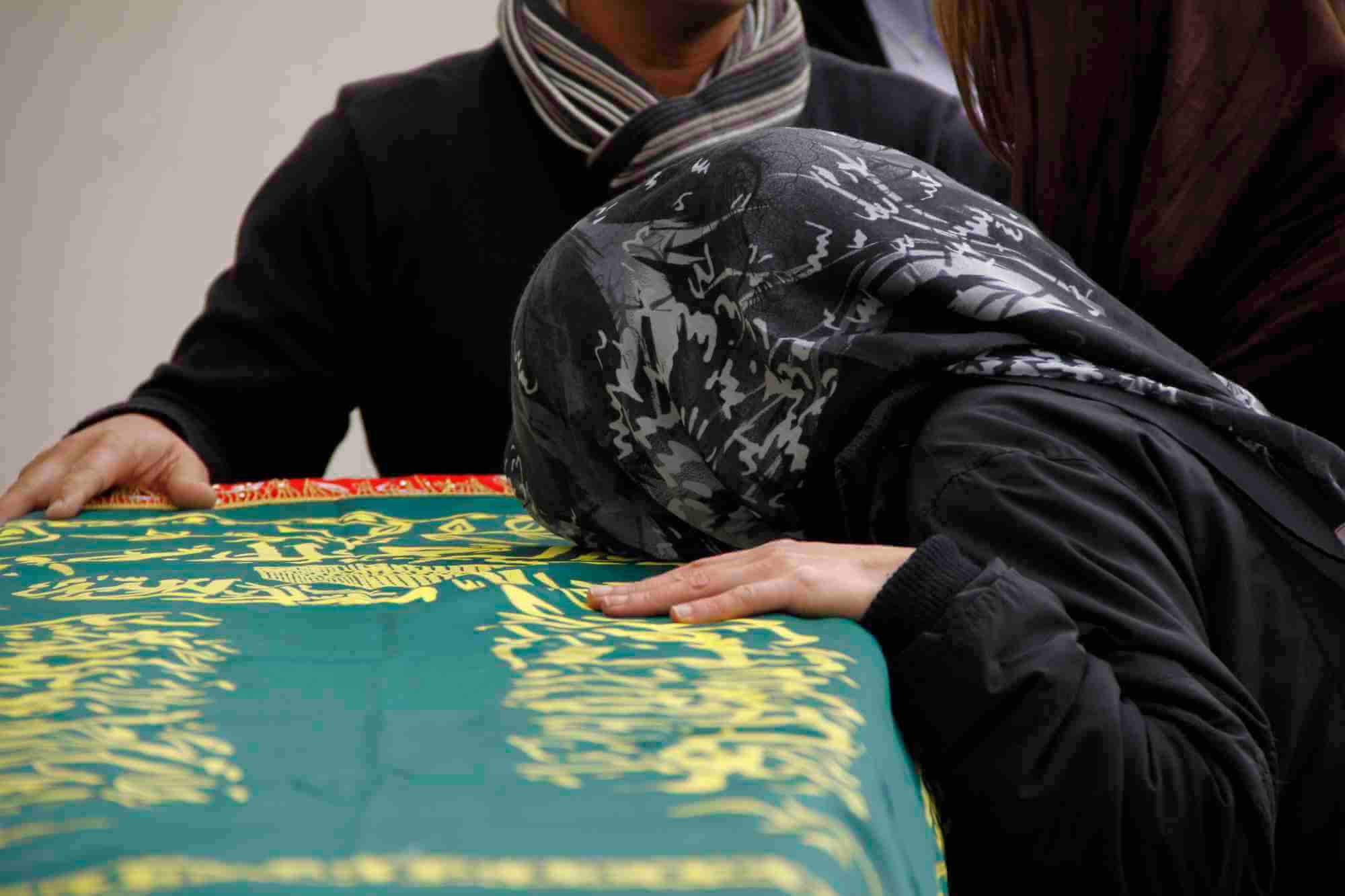 grieving muslim woman resting her head on a casket with arabic writing on it